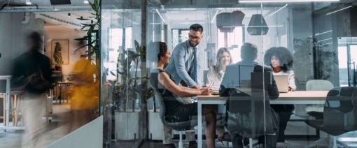 A group of five people sit and stand around a conference table in a modern glass-walled office, smiling and having a discussion, while two blurred figures walk by outside the room.