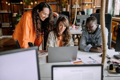 Three people happily collaborate around a laptop in a modern office setting.