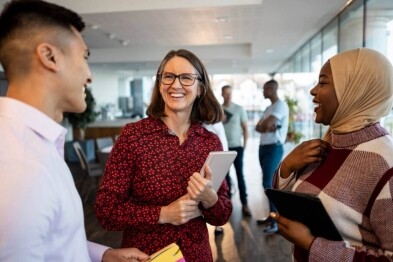 Three people are talking and laughing in an office setting. Others are in the background.