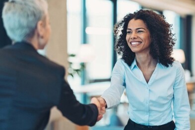 Two women shaking hands in a bright office, one smiling in a light blue shirt, the other with short gray hair