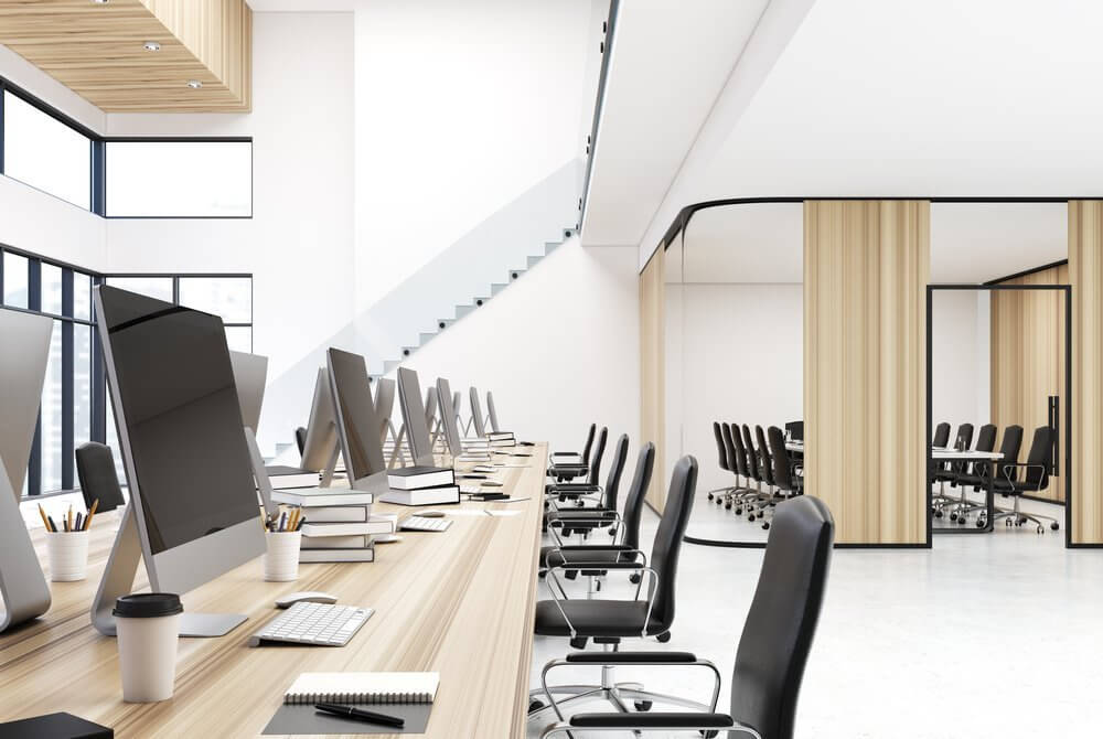 Modern office with rows of desks, computers, notebooks, and a conference room with chairs and a table in the background.