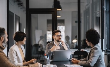 Four people having a discussion at a conference table in an office setting with laptops and documents.