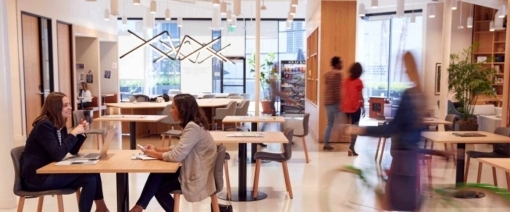 Modern office with people sitting at tables, having discussions—some even engaged in hybrid workplace platform comparison as part of their collaboration. Others walk or ride a bike through the space. Large windows, wooden accents, and shelves create a bright, open, and collaborative atmosphere.